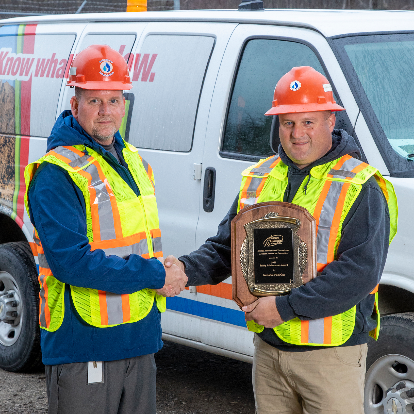 two men shaking hands holding an award in front of utility truck